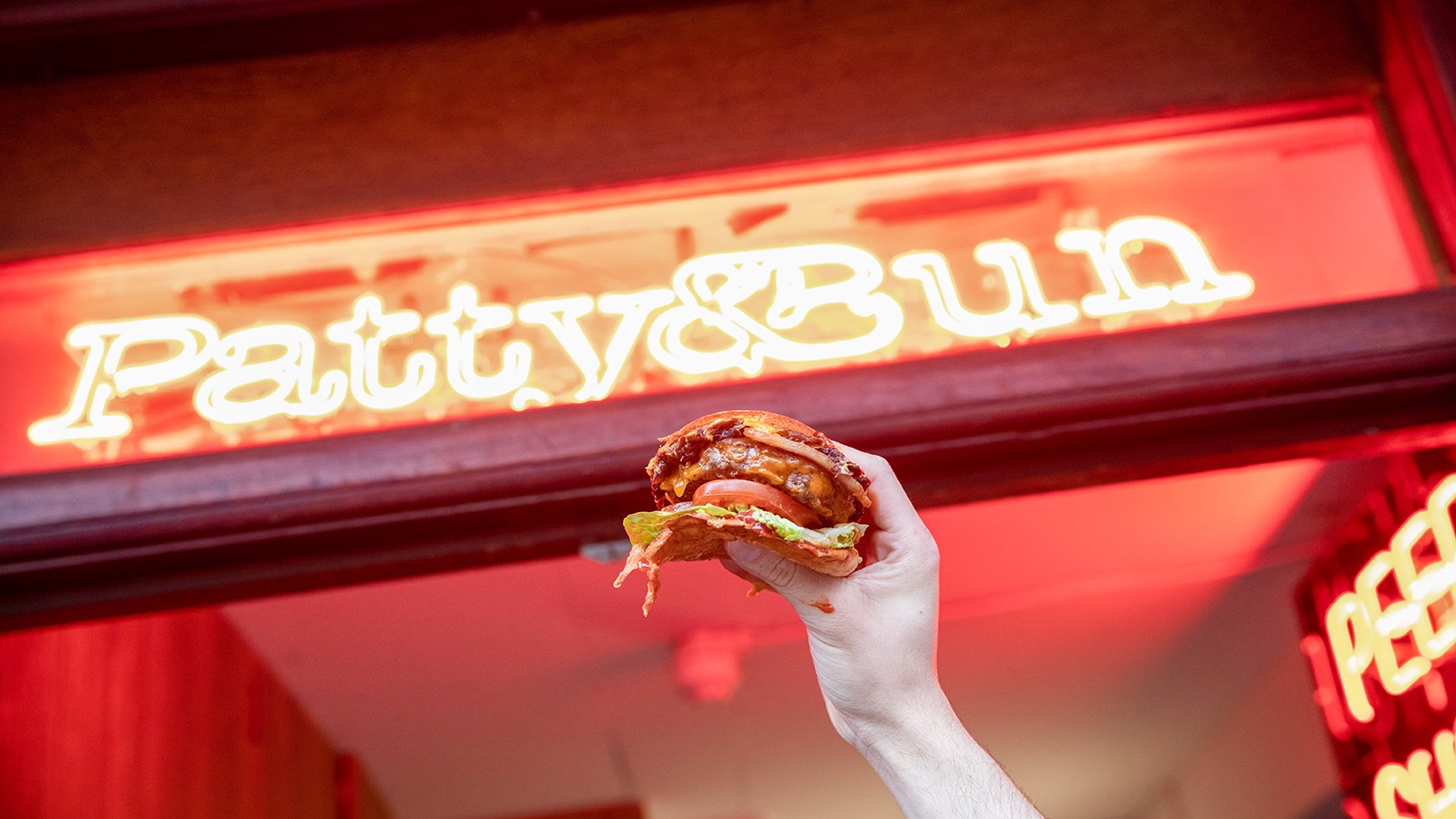 Patty & Bun burger held up beneath neon sign